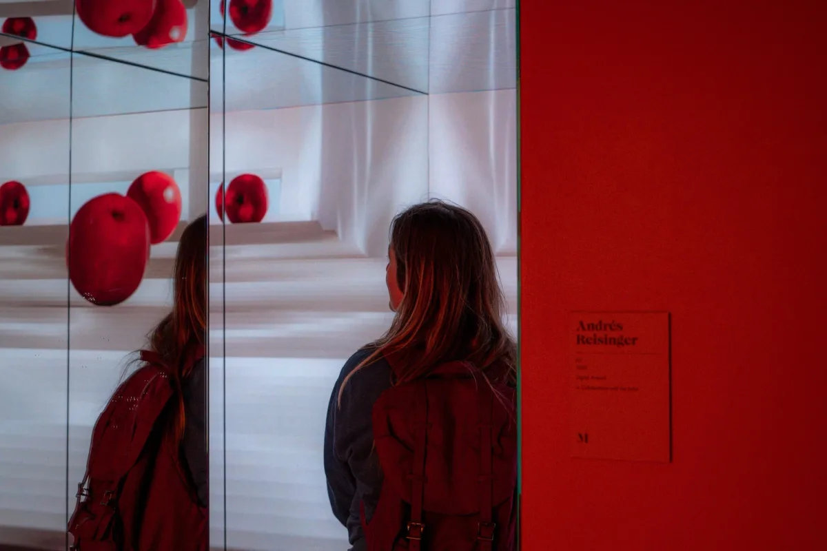 A woman standing in front of a mirror at an art gallery, observing a red apple sculpture reflected in the mirror. The gallery has a sign that reads 'Andrés Reisinger' on a red wall. Parham Motamedifard
