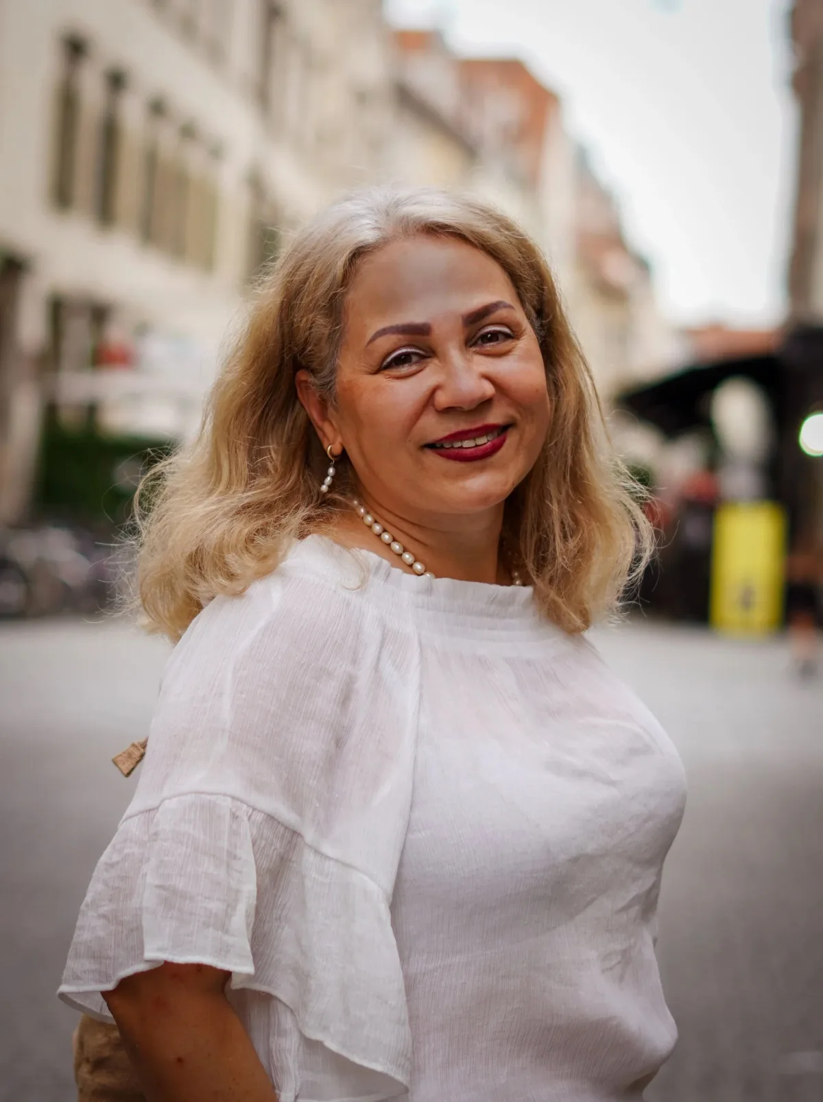 A smiling woman with blonde hair, wearing a white blouse, pearl necklace, and earrings, standing on a city street with buildings and blurred surroundings in the background. Parham Motamedifard